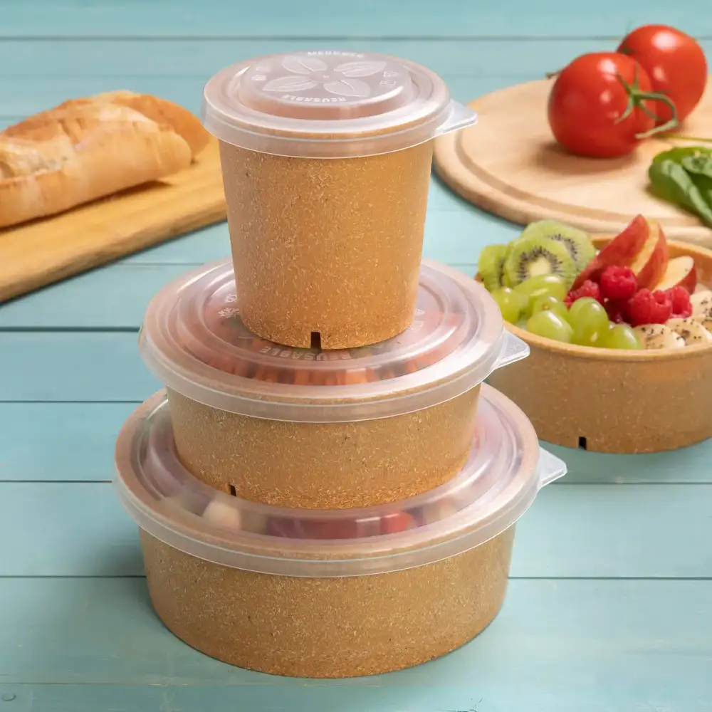 Empilement de trois conteneurs bruns avec des couvercles transparents sur une surface bleue, avec du pain, des tomates et une salade de fruits à proximité.