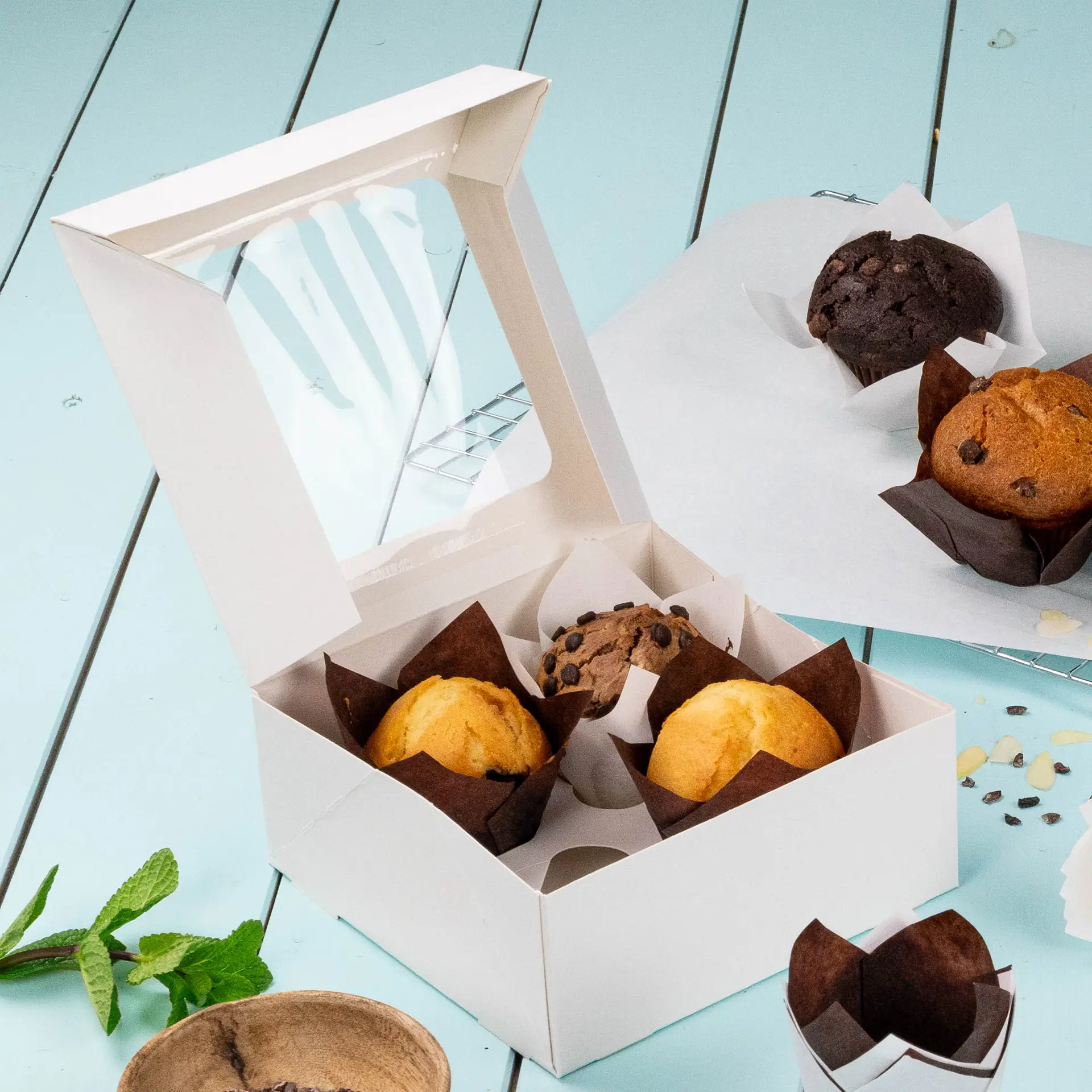 Boîte à cupcakes blanche de 4 pièces avec une fenêtre en cellulose, contenant des muffins et un muffin chocolat, sur une surface bleu clair.