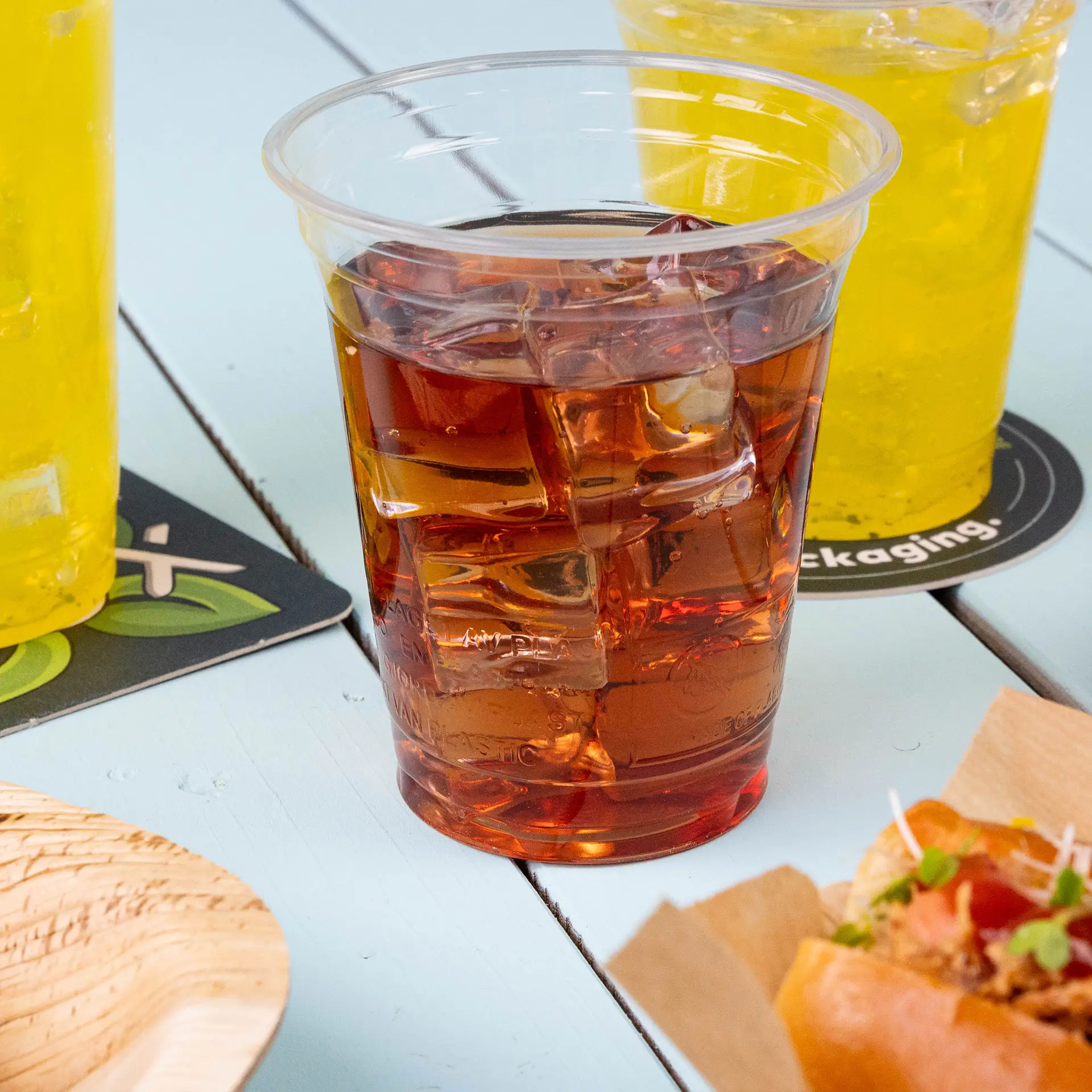 Gobelet en plastique transparent de 12 oz en bioplastique PLA rempli de thé glacé, entouré de boissons jaunes et d'un aliment.
